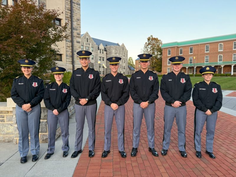 The seven cadets are all in uniform smiling wearing black jackets on Upper Quad with a historic brick building and a Hokie stone building in the background. 
