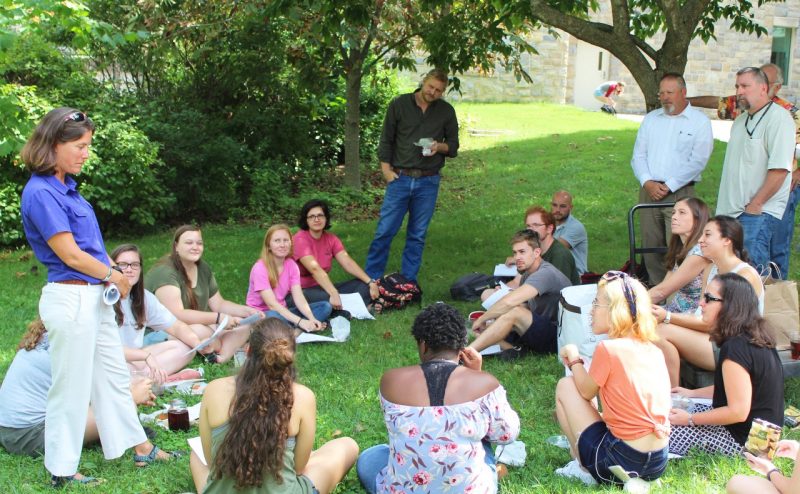 A professor standing speaks to a group of seated students, faculty, and staff outdoors under the shade of a tree.